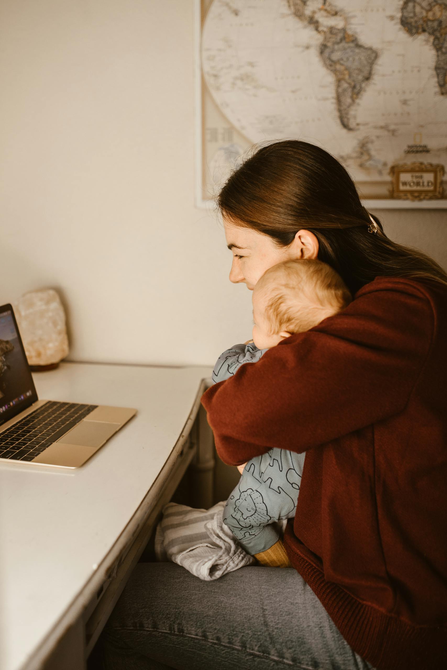A loving mother holds her baby while working on a laptop at home, capturing a tender moment.